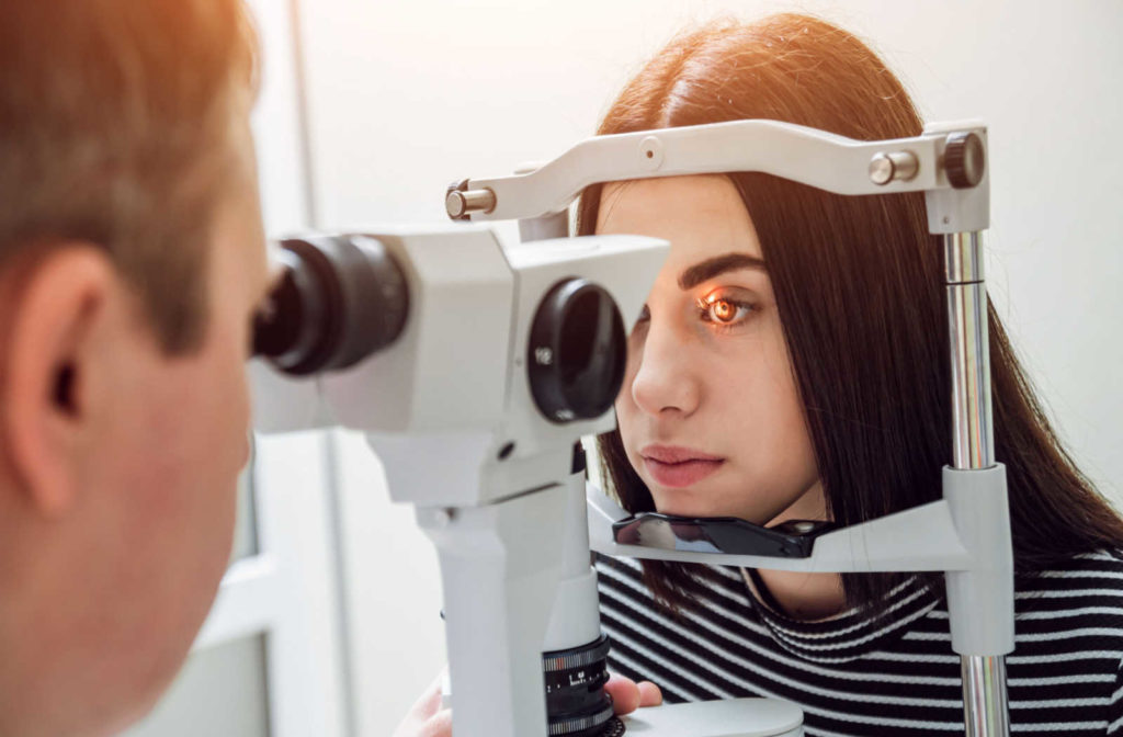 An optometrist performing a slit lamp test on a female patient with a light shining in her eye while she rests her head on the forehead and chin rest.