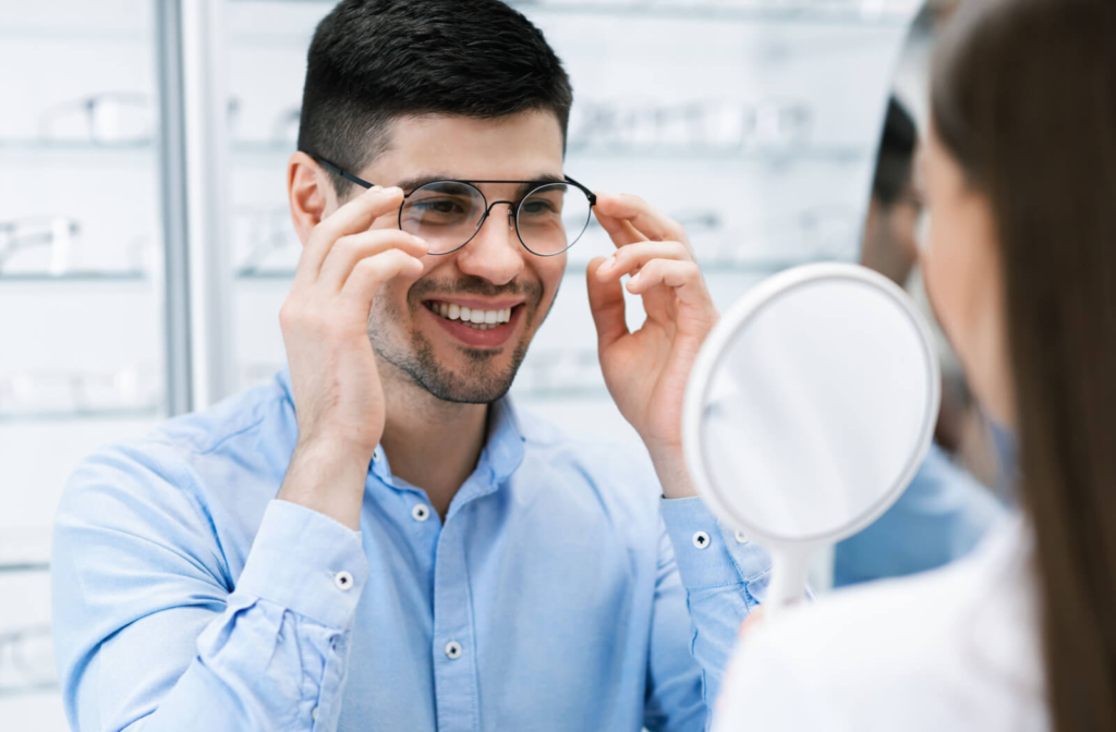 A young man trying on glasses at an optic store while being assisted by an optometrist.