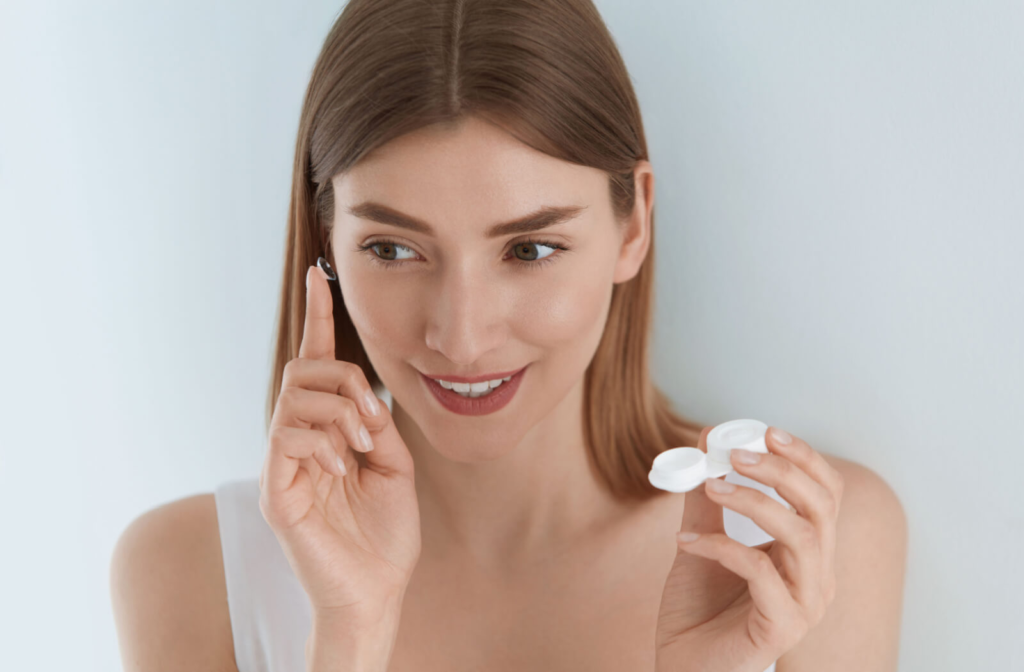 A woman smiling and holding a contact lens up to her right eye while holding a contact lens case in her left hand.