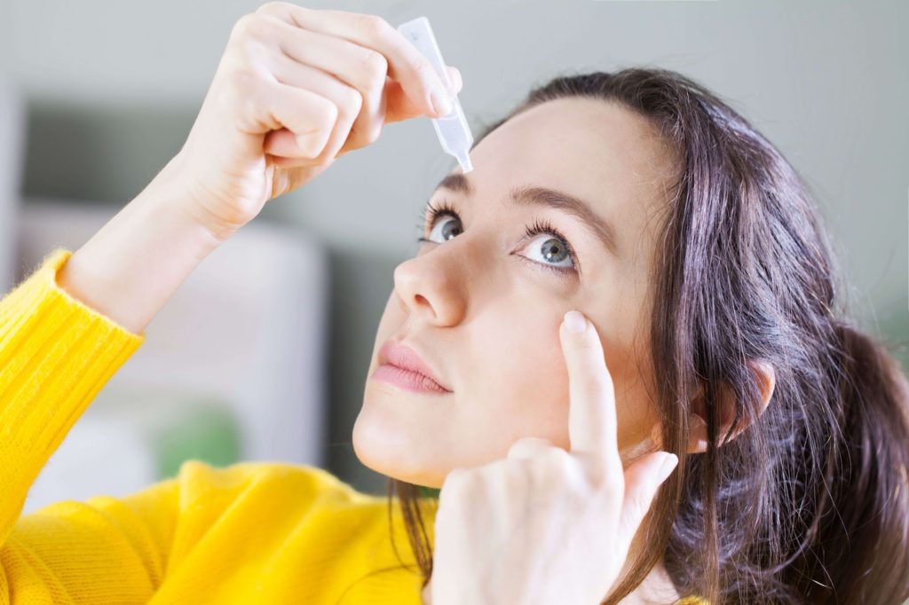 A woman holding a small bottle of eye drops in her right hand and putting them on her left eye while she uses her left index finger to pull her eyelid down.