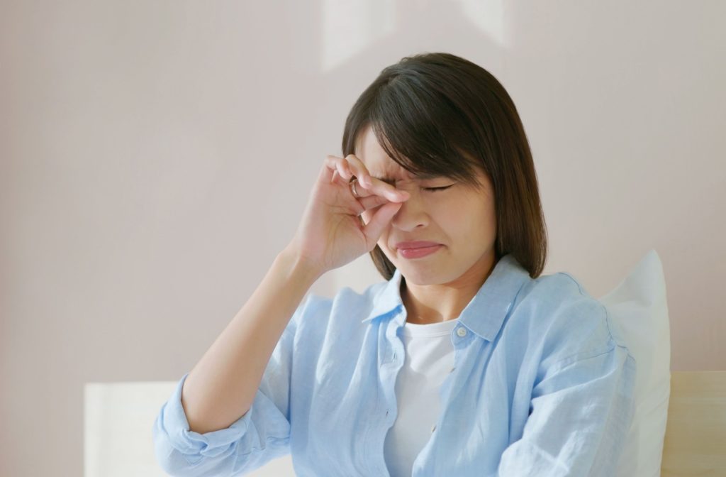 A woman sitting on a bed rubs her eyes to try and alleviate irritation from allergies.