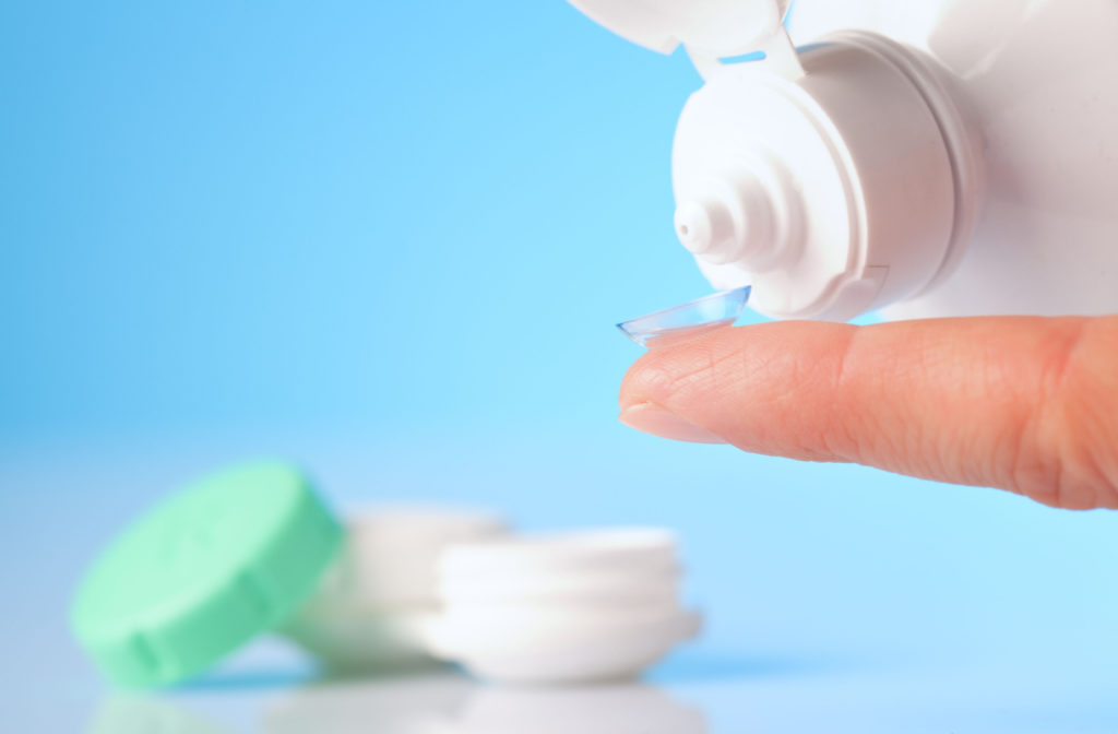 A close up of a contact lens on a finger tip, a bottle of contact lens solution being pouring onto the lens, and a contact lens case sitting in the background.