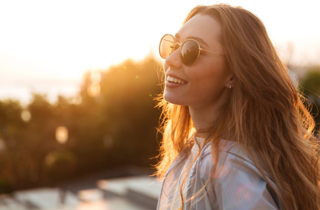 A woman outside, looking out into the distance, smiling with sunglasses on.