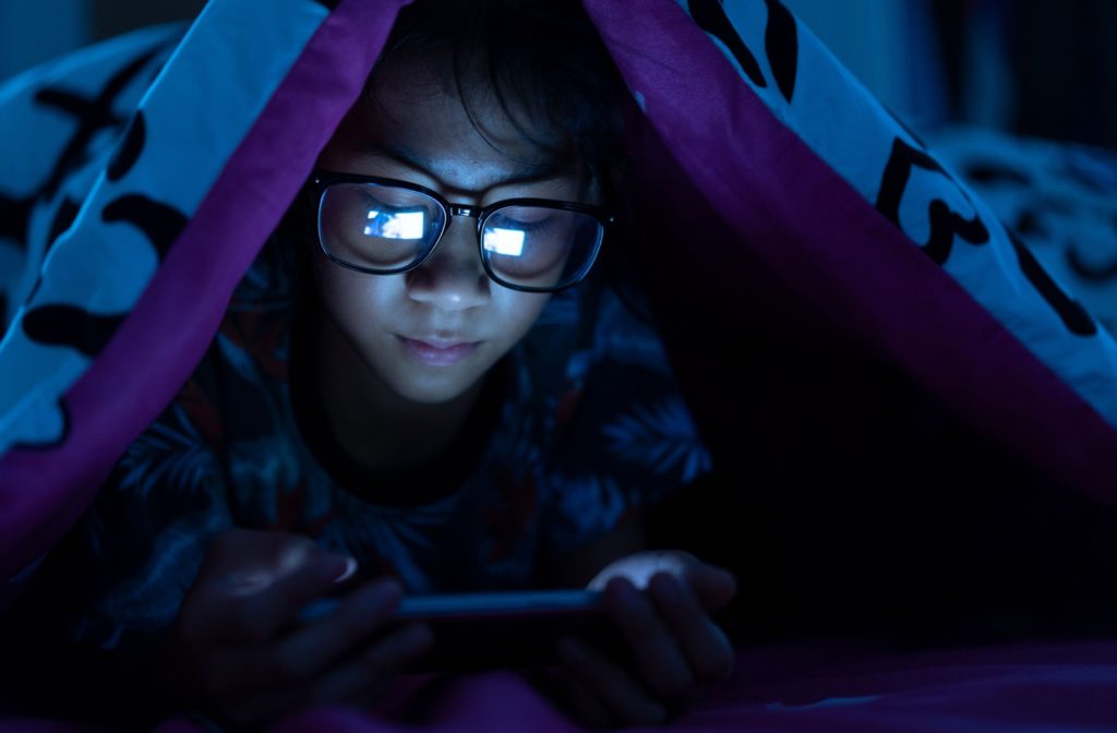 Young girl wearing blue light glasses while watching a video on her mobile phone in the dark.