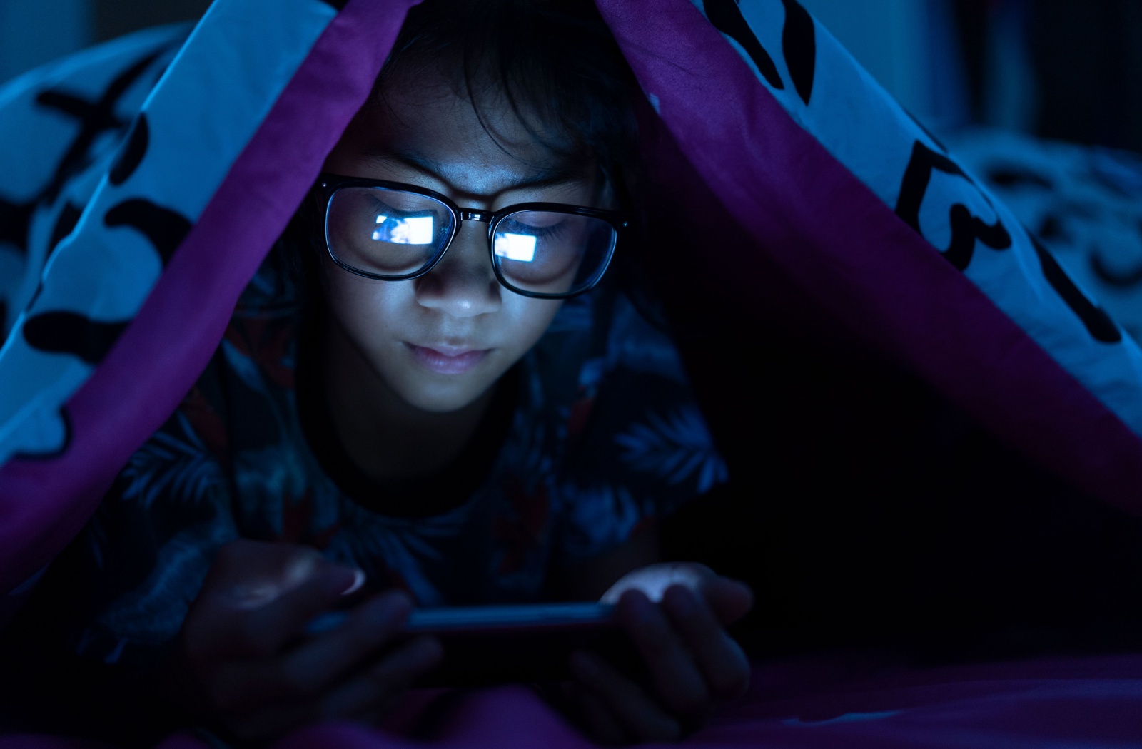 Young girl wearing blue light glasses while watching a video on her mobile phone in the dark.