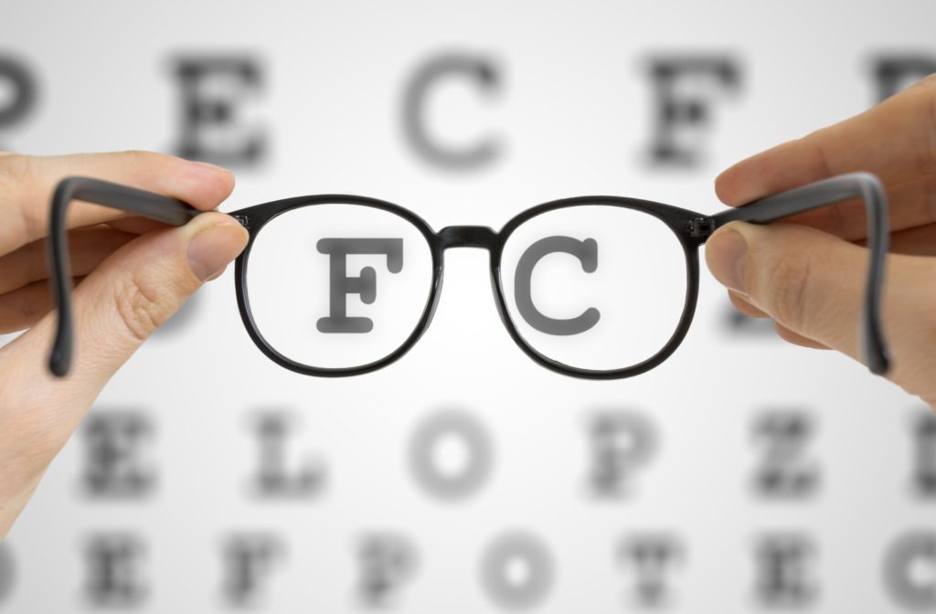 A pair of black framed glasses are held up against a blurred letter chart, with F and C appearing clearly in the lenses.