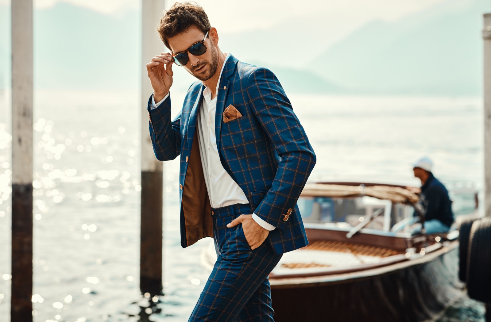 A person in a blue checkered suit walks along a dock with a boat in the background. They're holding their fashionable sunglasses while looking back over their shoulder.