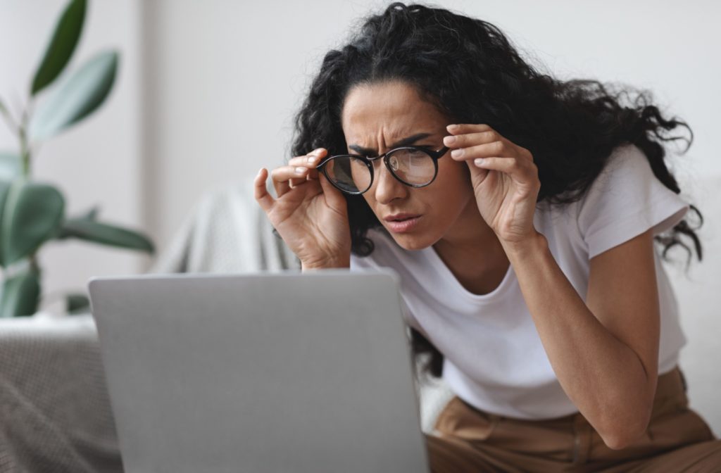 A person holds up their eyeglasses while squinting at their laptop screen.