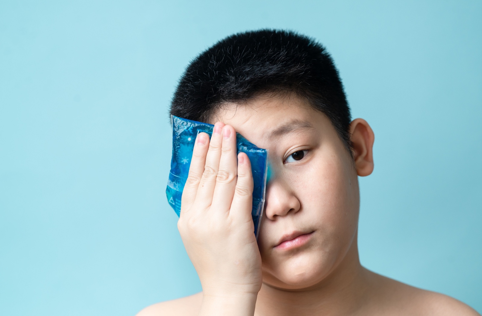 Against a pale blue background, a pre-teen holds a blue cold compress against a single eye to relieve swelling