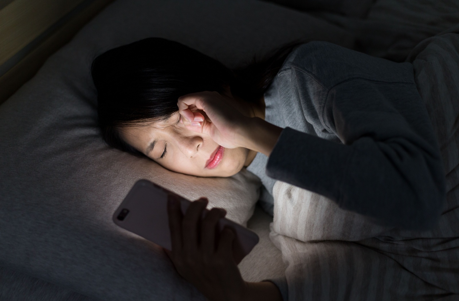 A person laying in bed, staring at a mobile phone, rubbing their eyes from extreme dry eye at night
