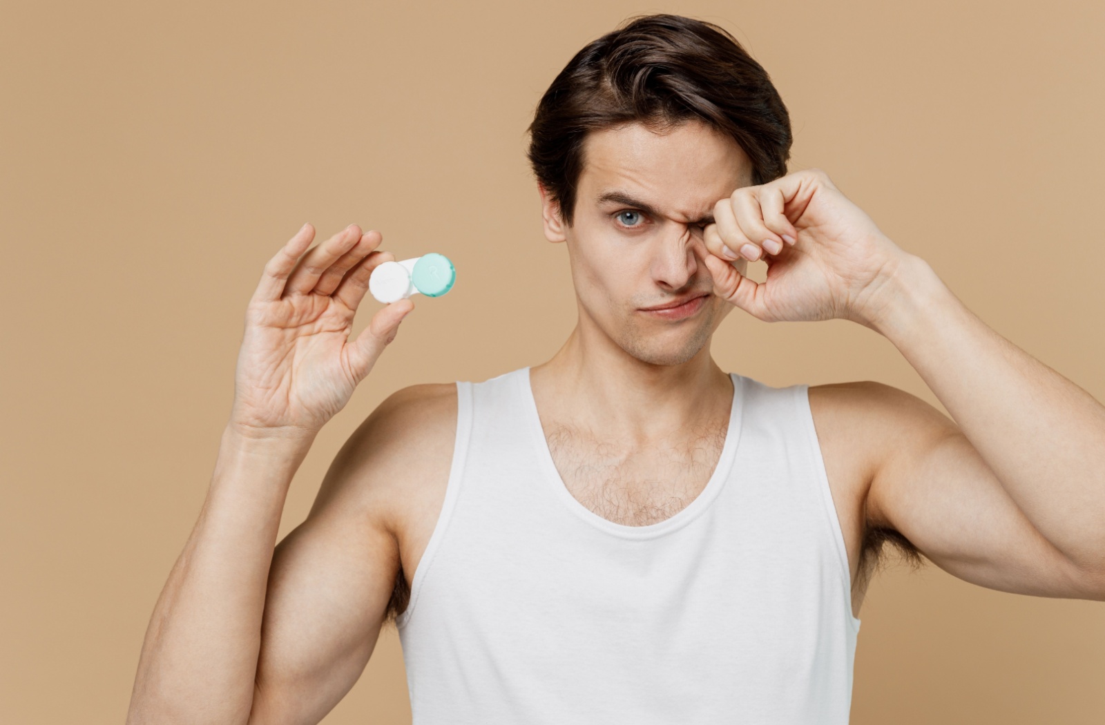 A person holds a contact lens storage case in one hand while rubbing their irritated eye with the other.