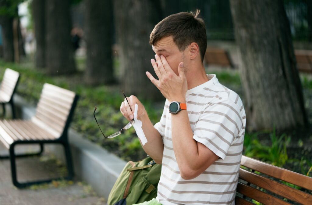 A person sitting on a bench outside holding their glasses in one hand and rubbing their eye with the other hand.