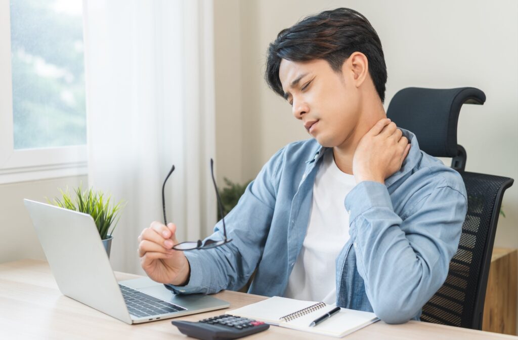 A person sitting at a desk, rubbing their neck due to pain related to eye strain and vision problems.